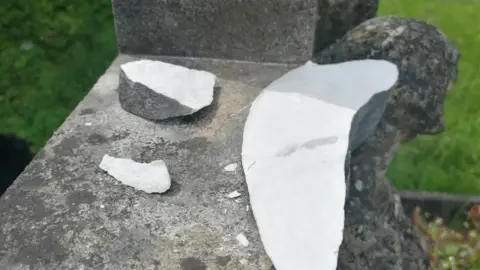 Arnos Vale Cemetery Trust A close up image showing the damage to an angel statue on top of a grave. The grey stone wing has been snapped, showing the white inside, and several shards lay beside it on the cross.