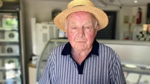 Frank looks at the camera with a small smile. He wears a grey, blue and white striped polo and a straw hat, and is in front of the counters full of ice cream inside the shop.