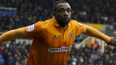 A footballer with dark hair, wearing a Wolves top, has his arms outstretched. Fans in a stand are blurred behind him.