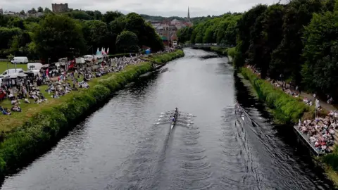 An aerial view of the Durham Regatta. Two rowing boats are on the River with corwds of people watching from the banks. The skyline of Durham City is in the background.