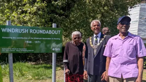 (l-r) Dorothy Grace Brown - wearing sunglasses and a black blouse holding crutches, next to the Mayor of Reading Glenn Dennis who is in a navy jacket with medals and Peter Small who is wearing a purple short sleeve shirt at the Windrush Roundabout