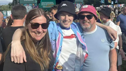 Family handout Owen and his parents are standing close together with their arms around each other at Tramlines Festival. Behind them, a large stage dominates the background, featuring banners and screens with text. Surrounding the stage is a crowd of people enjoying the event.