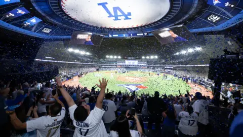 Cosm Inside a Cosm venue showing fans of the LA dodgers celebrating the 2025 World Series win. The screen shows a giant view of the pitch.