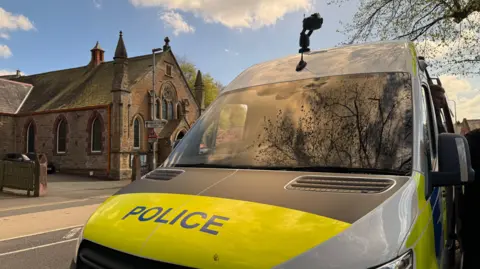 Close up photo of police van with a camera on top of it. The van is front of a church on a sunny day. The van says the words "police" in the wrong on a yellow background. 