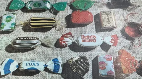 Gainsborough Heritage Centre More than a dozen wrapped sweets arranged in rows on a cream cloth. They include Fox's mints, with a white and blue wrapper, Nuttell's Mintoes (white and red), Keiller Butterscotch (gold wrapper), and another wrapped to look like a red strawberry with a green top. 