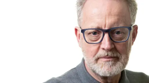 Simon Nicholls Man with grey hair and glasses and a beard looking directly at the camera. He is wearing a grey shirt against a white background.