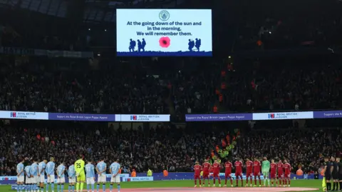 Reuters Manchester City and Liverpool teams line up for a minute's silence around the centre of the pitch with packed stands behind them. A big screen above says "At the going down of the sun and in the morning, we will remember them."
