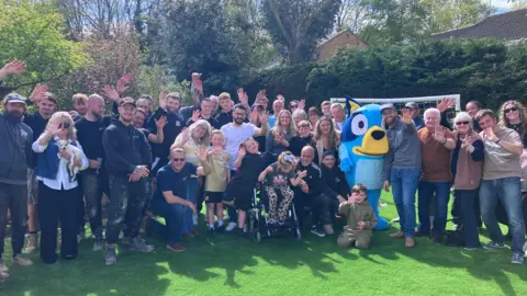 Ada and her family with all the volunteers and Swindon Town foorball team.
They are sitting and standing in the new garden with a 'Bluey' character, with trees behind them.
