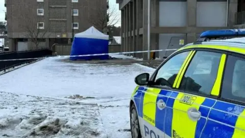 Police car at the scene of an accident, with snow on the ground and a blue police tent behind police tape, in the middle of apartment blocks