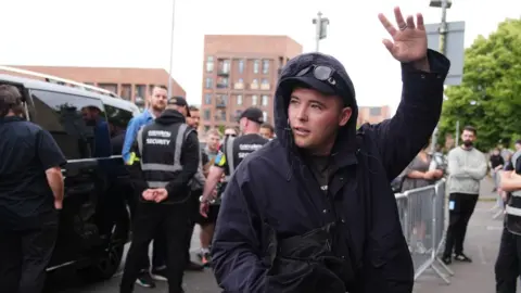 Liam Ó hAnnaidh, who is wearing a dark raincoat with the hood up over a black cap and sunglasses, waves to crowds outside Glasgow's O2 Academy. Behind him are his security team standing in front of a dark vehicle.