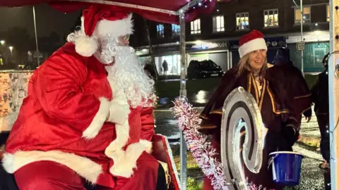 A man is dressed in a red Santa suit and is wearing a Santa hat. He has a long grey curly beard and is sitting on a sleigh. A woman is standing next to Santa wearing a red robe and holding a blue bucket.