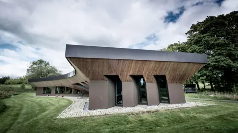 Maggie's Centres Maggie's Forth Valley centre. It is brown and made of stone and timber. The design is a boathouse with a timber umbrella connecting the wall to the roof. There is green grass around it and some trees in the background.