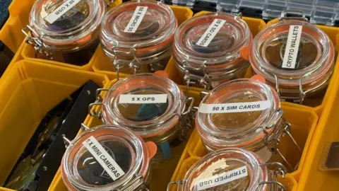 Kevin Shoesmith/BBC A yellow plastic box divided up into segments. Inside each segment is a glass Kilner jar containing digital items such as USB sticks, mini cameras and SIM cards for mobile phones.