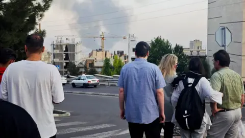People stand with their backs to the camera as they look at smoke rising in the horizon in Tehran on Monday