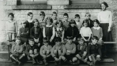 Orkney Library and Archive A black and white school photo from the 1930s. Three rows of 22 children. Robert Shaw is standing slightly apart from his classmates on the top left, wearing shorts and a light v-neck jumper with a pattern around the neck. A female teacher in a light top and dark skirt is standing at the other end of the rows. They are against a stone wall.