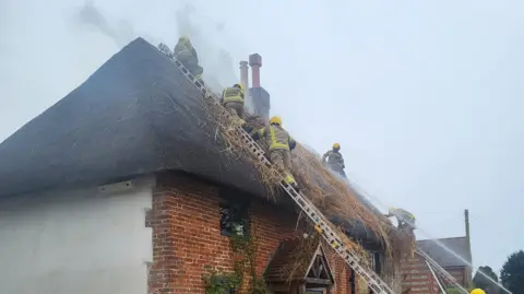 Firefighters working to extinguish a fire at a thatched cottage. There are several crew members climbing a ladder up to the roof. They are all wearing uniform and high vis helmets. There is grey smoke rising from the roof of the property and water from a hose can be seen from the bottom of the frame. 