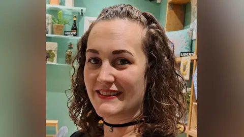 Royah Irvine Royah Irvine smiles at the camera. She has short curly hair, some of which has been pinned back. She wears a black choker necklace and a black top.