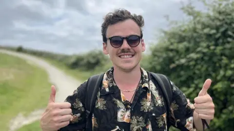 Woody Cook wearing a flowery shirt smiling at the camera with his thumbs up