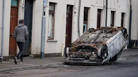 A man wearing a grey hoodie and tracksuit bottoms walks past an overturned and burnt out car on the street. There are a row of white terrace houses in the distance.