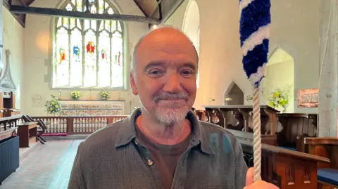 George Carden/BBC Guy Pratt stood in a cotton overshirt holding a rope connected to one of the church bells. He has a large stain glass window and the altar behind him