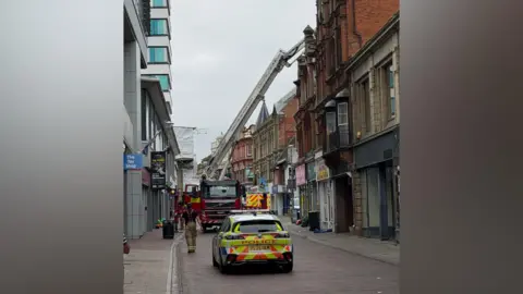 A police car, fire engine and ambulance are parked on a pedestrian street. A firefighter in blue t-shirt with yellow firefighting trousers is walking on the left hand side of the road. There is a crane appliance extended up to a roof.
