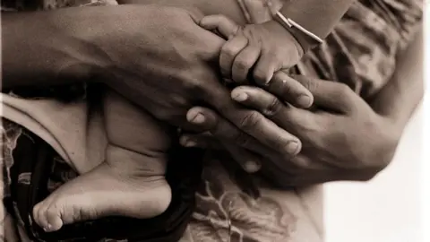 Fairfax Media via Getty Images A sepia-toned image of a baby's foot and hand being cuddled by an adult.