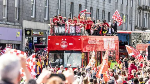 SNS Fans crowd around a red bus with football players on top holding a trophy