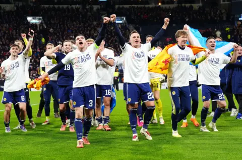 PA Media Scotland players celebrate the victory over Denmark on the pitch at Hampden. They are wearing white "We'll Be Coming" celebration t-shirts and waving Scotland flags.