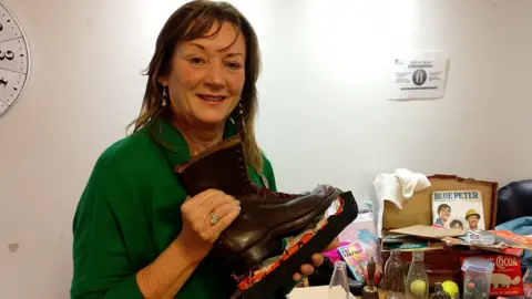 Sandra Leckey, in green sweater, holding up brown boots