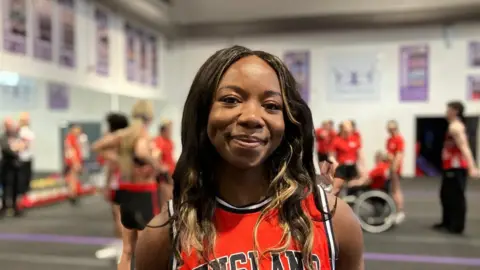 Tifé wears a red England training vest with a black and white outline. She has long dark brunette hair and stands in front of a training session taking place on the gym floor