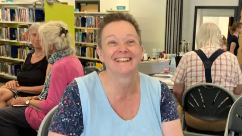 Jean Fenton is an older woman who is sat wearing a light blue pinafore over a dark blue flower patterned tee shirt.  She is wearing hearing aids in both ears.  She has short light brown hair and is smiling.