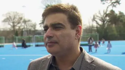 Muhammad Atif is looking off to the left. He has brown hair and is wearing a brown shirt and jacket. The backdrop is blue astro turf, and there are people and goals on the pitch.