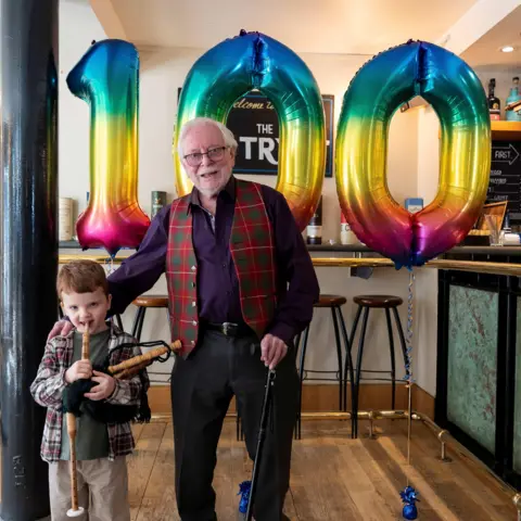 Hannah Power - National Piping Centre Bob Mcfie stands next to a small child holding what appears to be a miniature bagpipe. Behind them are large rainbow‑coloured balloons forming the number “100,”