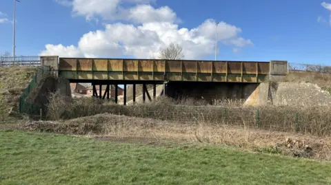 The ageing Winterstoke Road Bridge, which is a yellow and green metal structure with heavy rust damage. It stretches over a railway track beside some fields and houses on the other side.
