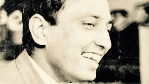 Jagwant Johal A side profile of Avtar Singh Jouhl in black and white. He is smiling with mouth open while looking to the right of the screen. He has short black hair and looks to be wearing a suit jacket, the collar shows, over a lighter top.