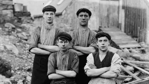 Four young men dressed in work wear, posed in front of the camera. They are wearing flat caps, shirts and aprons. 