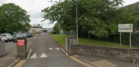 Google A general view from the road of St Joseph's Roman Catholic Comprehensive School in Aberavon. A gate is open leading into the school grounds with a waist-height stone wall surrounding the school boundary. A white sign with the school name on can be seen in the left hand side. Through the gate is a car park filled with cars with the school buildings in the background. 