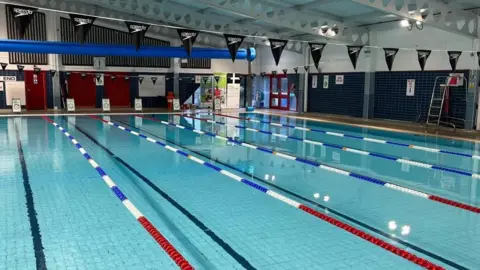 A picture of helston sports centre. It is a large swimming pool.  There are a number of lanes and bunting is hanging from the ceiling.