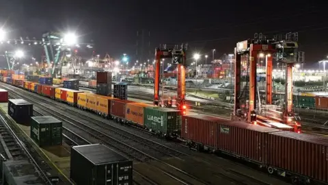 BBC Southampton Docks at night with a freight train loaded with containers in the foreground extends back into the distance. Floodlights illuminate stacks of freight containers are piled up in the background.