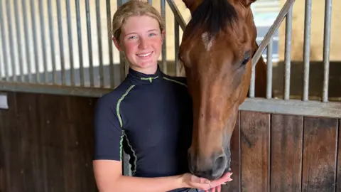 Racehorse trainer Lauren Frost in a dark riding top alongside one of the horses at her yard