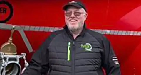 A man in a black jacket and black baseball cap is standing in front of a red background. 