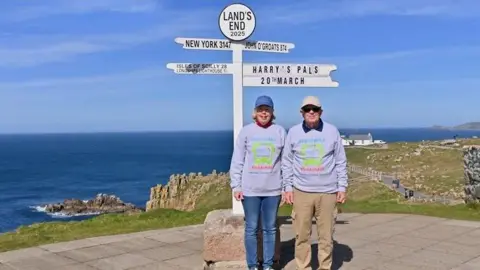 The Banhams next to the sign at Land's End with the sea behind them. The sign has directions to New York and John O'Groats, as well as a sign saying "Harry's Pals, 20th March". Mrs Banham is wearing a blue cap and a grey sweatshirt with blue jeans, while Mr Banham has a white cap, a grey sweatshirt and brown trousers. They are standing under the sign.