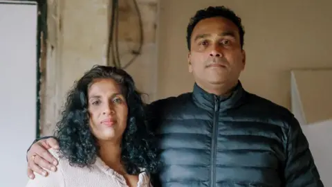 Don Hettiarachchi (right) a man with short dark hair wearing a dark padded jacket, standing next to his wife who has long dark hair and is wearing a pink cardigan. They are standing inside his burnt-out home, which has charred door frames and walls and stripped-out electrics