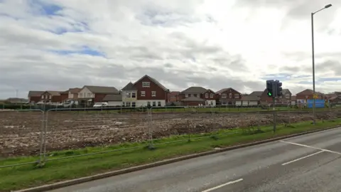 Google Maps A road with traffic lights and a new housing development site with barriers and houses set back off the road. 