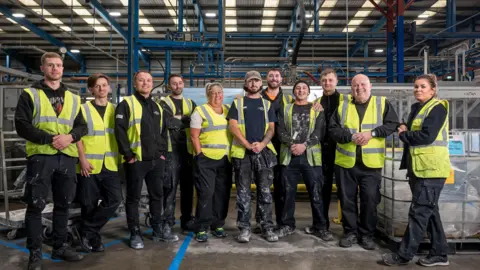 Group picture of 11 workers in hi-vis yellow jackets lined up on factory floor with blue metal frames behind them 