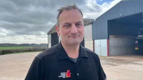 Ellen Knight/BBC Photo of Williamson looking into the camera and smiling slightly. He is wearing a black polo shirt with a red cow embroidered on the chest, and the words 'Nuffield Farming Scholarship Trust' embroidered in white lettering. He has short brown hair, and is stood in front of two large, corrugated iron barns. In one of the barns, you can see the corner of a large tractor. Beyond the barns are hills, with green fields and trees lining the horizon. The sky has patches of blue, with grey clouds and a bit of sunshine. 