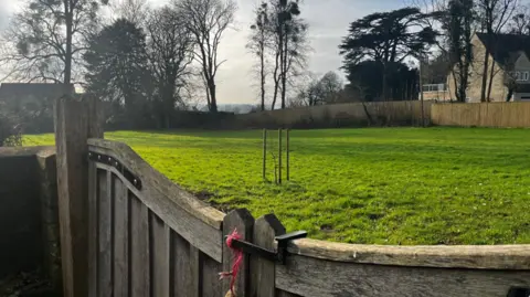 A shot across the top of a small wooden gate towards a green empty field with trees and a house in the background