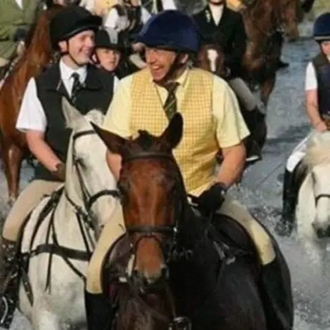 Nenthorn Equestrian Centre A man in helmet and tattersall waistcoat is riding a brown horse in a crowd of other riders. He is smiling broadly as they ride through water.