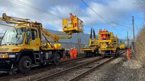 Workman wearing orange suits standing on cherry pickers, which are running along the Tyne and Wear Metro line. The staff are examining the overhead lines.