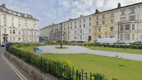 The Crescent in Bridlington. It shows a green area surrounded by black railings with pavement in the middle of the grass. A road surrounds the green area and there are residential flats.